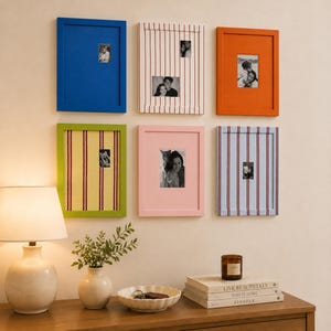 May include: Six rectangular wall-mounted picture frames arranged in two rows. The top row features a solid blue frame, a white frame with vertical red stripes, and a solid orange frame. The bottom row includes a lime green frame with vertical red stripes, a solid pink frame, and a light blue frame with vertical red stripes. Each frame contains a small black and white photograph.