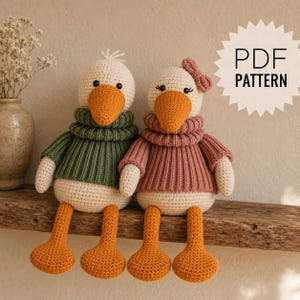May include: Two crocheted goose dolls with orange beaks and feet. One wears a green ribbed sweater, the other a pink ribbed sweater with a matching bow. The text PDF PATTERN is displayed in a white starburst shape on the right side of the image.