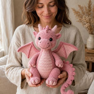 May include: A handmade crochet dragon plush toy in shades of pink. The creature features a rounded head with small horns, large black safety eyes, a stitched smile, two pointed wings, and a long tail with a row of small spikes along the back.