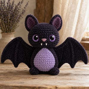 May include: A dark purple and light lavender crocheted bat plush toy featuring large glossy black eyes, a small pink nose, and two white fangs. The bat has pointed ears with lavender inner details and wide, outstretched wings made of dark yarn.