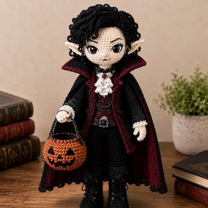 May include: A crocheted doll dressed as a vampire with black curly hair, pointed ears, and fangs. The doll wears a black and dark red cape, a white ruffled cravat with a red gem, and black boots. It holds a small, round, orange crocheted pumpkin basket.