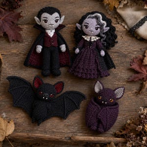 May include: Two crocheted vampire dolls and two crocheted bats. One vampire wears a black cape with a dark red lining, a vest, and trousers. The other wears a dark purple dress with a white lace collar. One bat is black, the other is purple.
