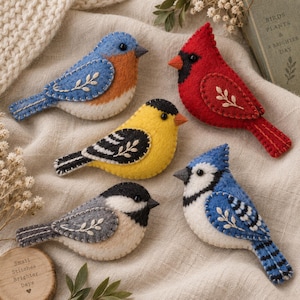 May include: Five handcrafted felt bird ornaments featuring embroidered leaf details and black bead eyes. The collection includes a bluebird, a red cardinal, a yellow goldfinch, a gray chickadee, and a blue jay, each with visible blanket stitching.