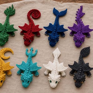 Op de afbeelding: Acht gehaakte drakenfiguren gerangschikt in twee rijen. De draken zijn groen, rood, donkerblauw, paars, geel, taling, wit en zwart. Elk wezen heeft gestructureerde schubben, puntige vleugels en een unieke staartvorm, allemaal gemaakt van garen.