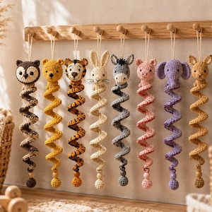 Op de afbeelding: Acht gehaakte dierenornamenten hangen aan een houten rek. De dieren zijn een uil, beer, tijger, konijn, ezel, varken, olifant en kangoeroe. Elk figuur heeft een lang, spiraalvormig lichaam dat eindigt in een kleine, ronde houten kraal.