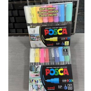 May include: Two sets of eight Uni Posca water-based paint markers in 0.7mm fine tip. The top set features soft pastel colours, while the bottom set contains standard colours. Each package displays the Posca logo, product details, and illustrations of decorated items.