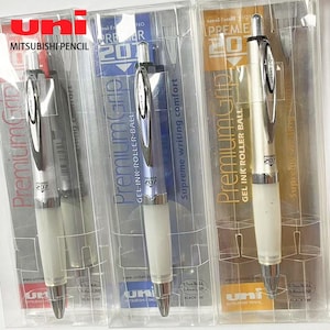 May include: Three Uni-ball Premier 207 gel ink rollerball pens in individual clear plastic packaging. Each pen features a silver-toned body, a white rubberized grip section, a metal pocket clip, and a 0.7mm ball tip for black ink writing.