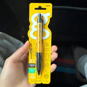 May include: A yellow and black Uni Alpha Gel Switch mechanical pencil in its original packaging. The packaging features the text Uni, Alpha Gel, Switch, Kuru Toga, and Japanese characters. The pencil is designed for 0.5 mm lead and includes a clip.