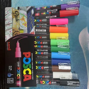 May include: A set of 12 Uni Posca PC-1M water-based paint markers in various colours, including purple, red, pink, orange, yellow, light green, green, light blue, blue, brown, white, and black, presented in a black cardboard display box.