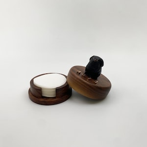 Op de afbeelding: Een ronde houten houder met een donkerbruine afwerking, met daarin een stapel witte ronde vellen papier. Het deksel is bekroond met een klein, uitgesneden zwart hondenfiguurtje en bevat vier kleine, metalen letterbedels die A-B-C-D spellen.