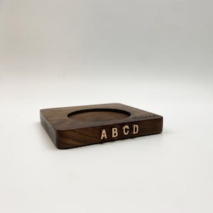 May include: A rectangular dark brown wooden coaster featuring a circular recessed center. The front edge displays the gold-colored letters A, B, C, and D, while the word mignon is engraved in the top right corner of the wooden surface.