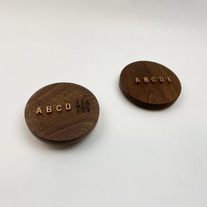 Könnte beinhalten: Zwei kreisförmige, dunkelbraune Holzscheiben mit sichtbarer Maserung. Die linke Scheibe zeigt die goldenen Buchstaben A B C D und eine kleine Grafik mit drei vertikalen Reglern. Die rechte Scheibe zeigt die goldenen Buchstaben A B C D E mittig.
