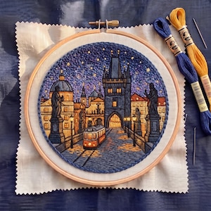 May include: A circular embroidery hoop containing a detailed needlework scene of a city bridge at night, featuring a tram, stone statues, and illuminated buildings under a starry sky. Two skeins of blue and yellow embroidery floss lie nearby.