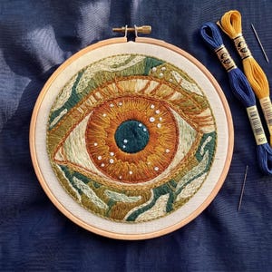 Könnte beinhalten: Ein Stickrahmen mit einem runden Stoffstück, das ein detailliertes, gesticktes Auge zeigt. Die Iris ist in Orange- und Brauntönen gehalten, mit einer dunkelblauen Pupille und weißen Akzenten. Zwei Stickgarnstränge in Blau und Gelb liegen daneben.