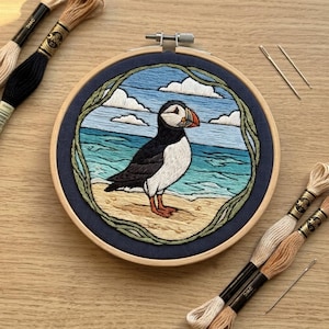May include: A circular wooden embroidery hoop containing a hand-stitched puffin on a sandy beach with a blue ocean and sky background. The design is framed by a green vine border. Several skeins of embroidery thread and two needles are included.