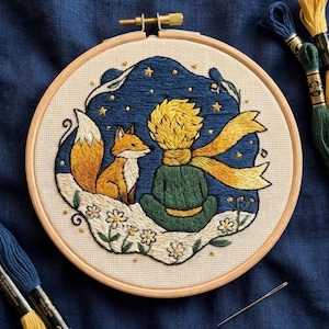Op de afbeelding: Een ronde houten borduurring met een handgestikt ontwerp van een persoon met geel haar en een groene outfit, zittend naast een oranje vos. De scène is geplaatst tegen een donkerblauwe sterrenhemel met witte bloemen op de grond.