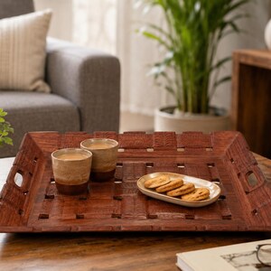 Pu&ograve; includere: Un vassoio da portata quadrato in legno marrone scuro con un motivo a intreccio e manici laterali integrati. Il vassoio contiene due tazze cilindriche in ceramica con una bevanda marrone chiaro e un piattino ovale con quattro biscotti.