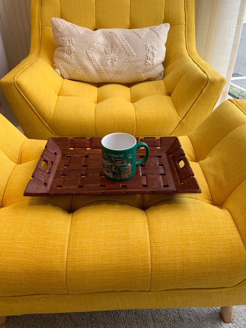 Pu&ograve; includere: Un vassoio rettangolare in legno marrone scuro con superficie intrecciata e manici intagliati, che sostiene una tazza in ceramica verde con la scritta "TURKIYE" e varie illustrazioni grafiche stampate, appoggiato su una poltrona giallo brillante.