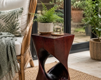 Può includere: Un tavolino in legno marrone scuro con una base ritorta a forma di clessidra e un piano quadrato piatto. Il tavolo presenta una finitura liscia e lucida con venature del legno visibili e una sezione centrale vuota, offrendo una base solida.