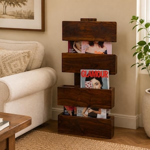 May include: A dark brown wooden magazine rack featuring four horizontal rectangular storage compartments stacked vertically. The rack holds several magazines, including one with the visible text Glamour, Daisy, and Big Fashion on the covers.