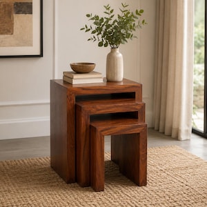 May include: A set of three nesting tables crafted from dark brown wood with a visible grain. The tables are stacked in a descending size order, featuring a clean, rectangular design. A ribbed ceramic vase with green foliage sits on top.