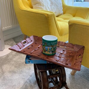 Pu&ograve; includere: Un vassoio rettangolare in legno con superficie intrecciata e manici intagliati, con sopra una tazza in ceramica verde decorata con una stampa grafica colorata di monumenti e la scritta T&Uuml;RKIYE, poggiato su un tavolino in legno scuro.