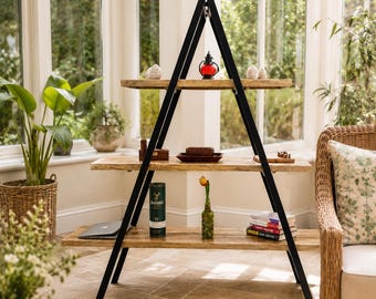 Può includere: Una scaffalatura a tre livelli a forma di A con una struttura in metallo nero e tre ripiani orizzontali in legno marrone chiaro. I ripiani ospitano vari oggetti decorativi, tra cui una lanterna rossa, una bottiglia verde, libri e piccoli contenitori.