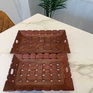 Pu&ograve; includere: Due vassoi rettangolari in legno marrone caratterizzati da un motivo a intreccio con dettagli testurizzati intagliati sulla superficie. Ogni vassoio presenta manici integrati sui lati corti e un design traforato su base e bordi.