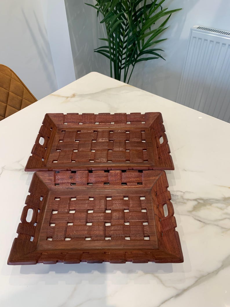 Pu&ograve; includere: Due vassoi rettangolari in legno marrone caratterizzati da un motivo a intreccio con dettagli testurizzati intagliati sulla superficie. Ogni vassoio presenta manici integrati sui lati corti e un design traforato su base e bordi.