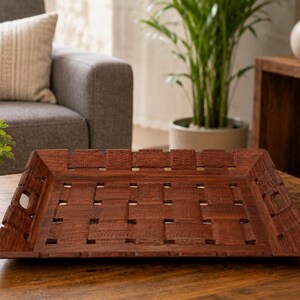 Pu&ograve; includere: Un vassoio rettangolare in legno con finitura marrone scuro e un design a intreccio. Il vassoio presenta una base piatta con spazi quadrati aperti tra le doghe intrecciate e manici ritagliati integrati su due lati opposti del bordo rialzato.