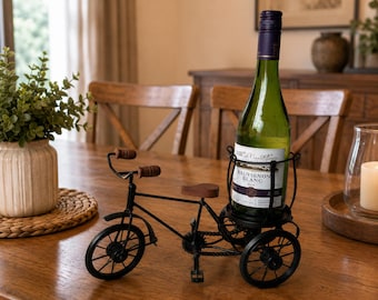 Può includere: Un portabottiglie da vino in metallo nero a forma di triciclo, con manubrio e sellino in legno marrone. Il telaio del triciclo include un supporto circolare sul retro progettato per sostenere una bottiglia di vino in posizione verticale.