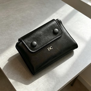 以下が含まれることがあります： 滑らかな質感の黒い財布で、2つの黒い円形スナップボタンで留める折りたたみ式のフラップが付いています。財布の前面には、フラップの下に金色の文字で「DC」と刻印されています。