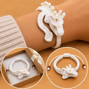 Könnte beinhalten: Ein weißes, gehäkeltes Drachenarmband mit kleinen goldfarbenen Perlen entlang des Rückens und Kopfes. Das Armband hat eine verstellbare Kordel mit zwei Holzperlen an den Enden. Es ist am Handgelenk sowie in einer Geschenkbox abgebildet.