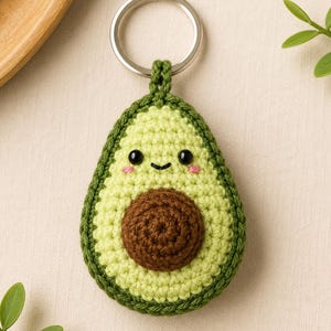Könnte beinhalten: Ein gehäkelter Avocado-Schlüsselanhänger mit dunkelgrünem Rand, hellgrünem Kernbereich und einem braunen Kern. Die Avocado hat zwei kleine schwarze Perlenaugen, ein aufgesticktes schwarzes Lächeln und zwei winzige rosa Wangen. Ein silberner Metallring ist befestigt.