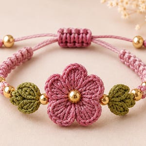 Könnte beinhalten: Ein Armband mit einer zentralen gehäkelten rosa Blume mit einer goldenen Perle in der Mitte, flankiert von zwei gehäkelten grünen Blättern. Das Band besteht aus rosa Kordel mit Goldperlen und einem verstellbaren Schiebeknotenverschluss.