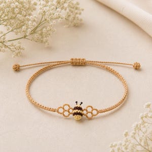 Könnte beinhalten: Ein beiges Makramee-Armband mit einem zentralen Bienenanhänger mit schwarzen und gelben Streifen, flankiert von zwei kleinen wabenförmigen Akzenten. Das Armband hat einen verstellbaren Schiebeknotenverschluss mit zwei kleinen geknoteten Enden.