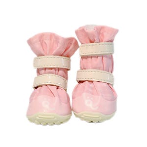 May include: A pair of light pink pet boots featuring a gathered fabric upper, two horizontal cream-colored hook-and-loop fastener straps, and a textured cream-colored rubber sole with a small white paw print logo on the front toe area.
