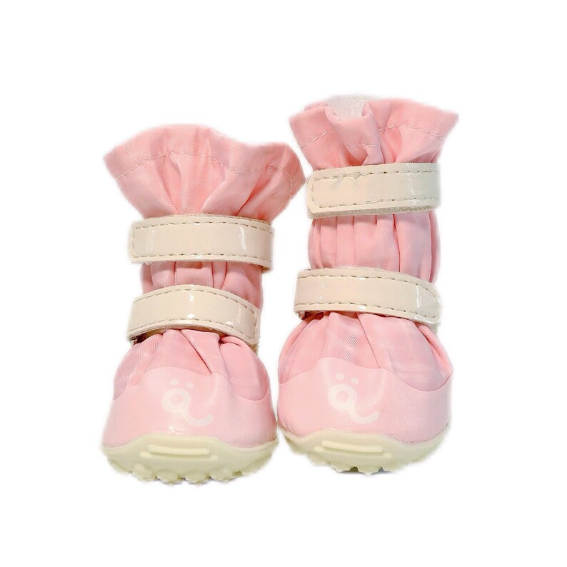 May include: A pair of light pink pet boots featuring a gathered fabric upper, two horizontal cream-colored hook-and-loop fastener straps, and a textured cream-colored rubber sole with a small white paw print logo on the front toe area.