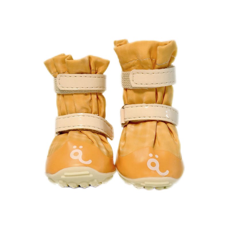 May include: A pair of mustard yellow pet boots featuring two beige hook-and-loop straps on each shaft. The boots have a rounded toe cap with a white graphic symbol resembling a stylized letter a with two dots above it and a textured sole.