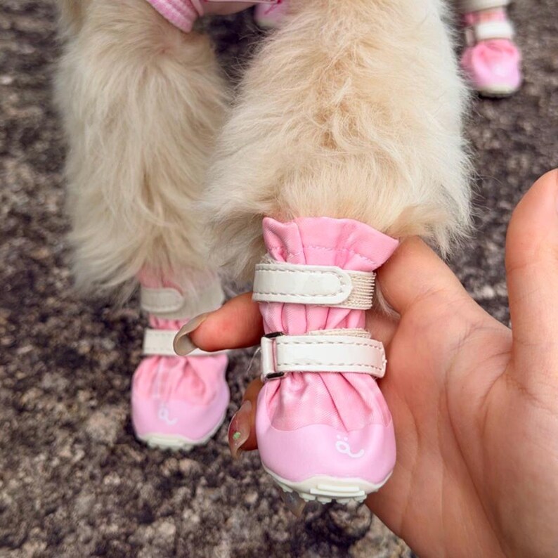 May include: A pair of light pink pet boots featuring a gathered fabric top, a reinforced toe cap, and two white adjustable hook-and-loop straps. The boots are designed for animal paws and include a small white graphic detail on the front toe area.