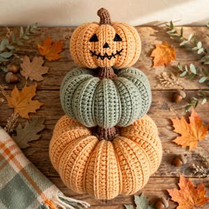 Puede incluir: Una pila de tres calabazas tejidas a ganchillo en tonos naranja y verde salvia. La calabaza superior presenta una cara de jack-o'-lantern bordada en negro. Cada calabaza tiene un tallo marrón tejido, mostrando un acabado de hilo texturizado.