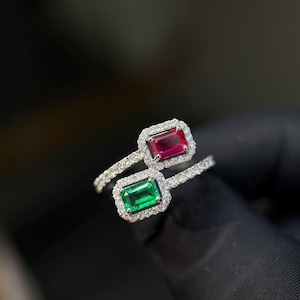 Könnte beinhalten: Ein Bypass-Ring mit zwei im Smaragdschliff geschliffenen Edelsteinen, einem leuchtend roten und einem tiefgrünen, die jeweils von einem Kranz aus kleinen weißen Steinen umgeben sind. Die Ringschiene besteht aus silberfarbenem Metall mit Besatz.