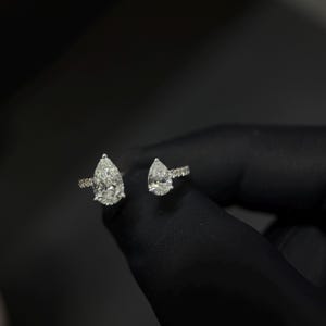 Könnte beinhalten: Ein offener Ring mit zwei birnenförmig geschliffenen Diamanten auf einer schmalen, mit kleinen Steinen besetzten Ringschiene. Der größere birnenförmige Diamant befindet sich auf einer Seite, während ein kleinerer Diamant am anderen Ende sitzt.
