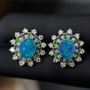 Könnte beinhalten: Ein Paar Ohrstecker mit einem zentralen ovalen blau-grünen Opal, umgeben von einer Anordnung aus runden weißen Diamanten, kleinen blauen Saphiren und grünen Smaragden, gefasst in einen gelbgoldenen Metallrahmen mit floralem Design.