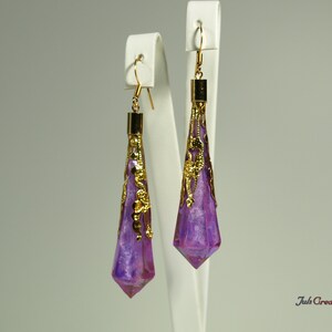 May include: A pair of long, purple and gold earrings with a pointed, faceted design. The earrings are hanging from white display stands.