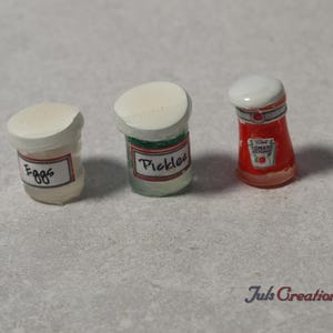 May include: Miniature food items including a jar of "Eggs", a jar of "Pickles", and a ketchup bottle. The jars have white lids and labels with handwritten text. The ketchup bottle is red with a white label.