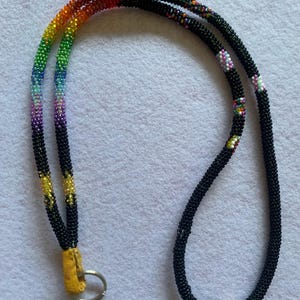 May include: A beaded lanyard featuring a continuous tube of small black glass beads. One side displays a vibrant rainbow gradient pattern, while the rest of the cord is primarily black with occasional small multi-coloured bead accents. A metal ring is attached to a yellow leather tab.