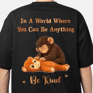 Pode incluir: Uma t-shirt preta com um gráfico de um macaco castanho a cuidar de um macaco de peluche cor de laranja. Por cima do gráfico está o texto In A World Where You Can Be Anything e por baixo o texto Be Kind com dois corações.