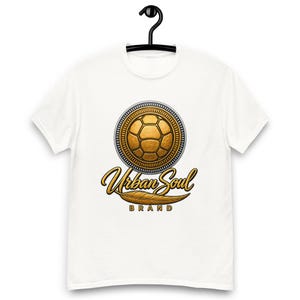 Op de afbeelding: Een wit t-shirt met korte mouwen en een centrale afbeelding van een gouden voetbal in een cirkelvormig kader. Onder de cirkel staat de tekst Urban Soul in een goudkleurig scriptlettertype, met het woord BRAND eronder en een gouden veer.