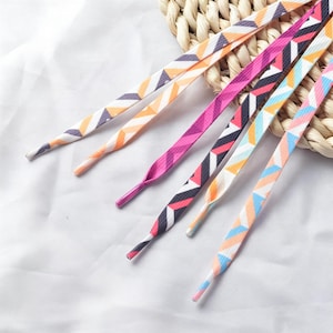 May include: A set of flat shoelaces featuring various geometric patterns and vibrant color combinations including orange, white, gray, pink, black, and light blue. Each lace has a solid colored plastic aglet at the tip for easy threading.